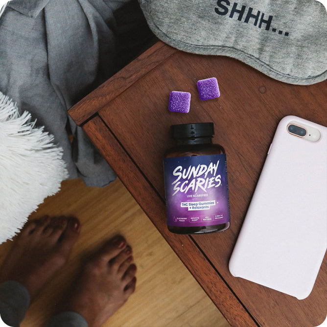 A person stands over their nightstand with an eye mask, cell phone and Sunday Scaries gummies on it.