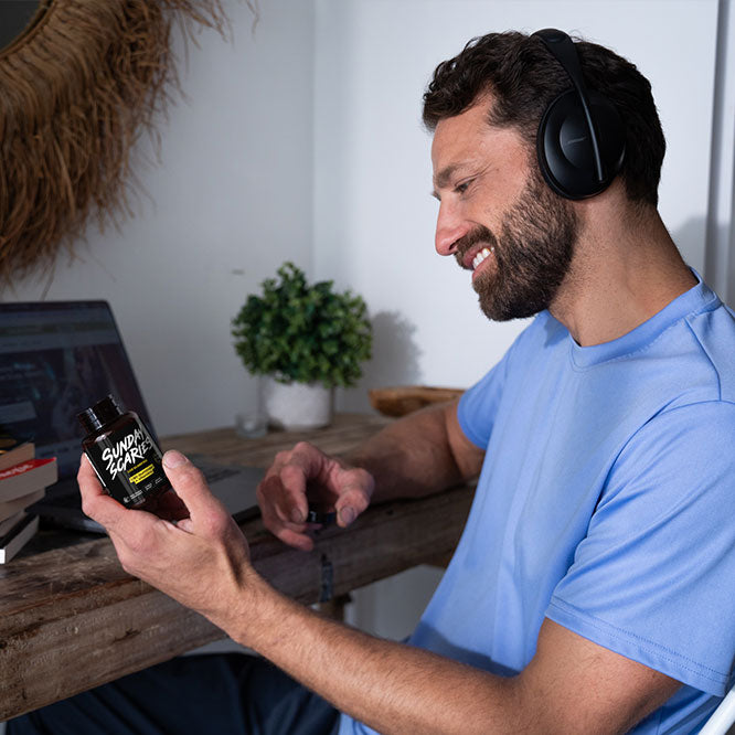 A man sits in front of his computer with his headphones on reading the back label of a Sunday Scaries gummy bottle