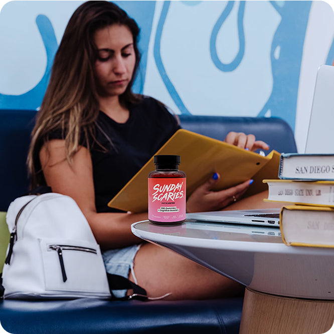 A woman studies at a library with books and a bottle of Sunday Scaries CBD focus gummies on the desk