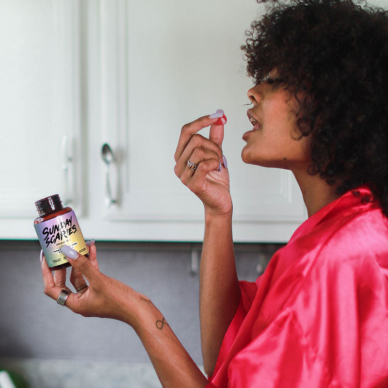A woman in her nightgown in a modern kitchen eats a delta-9 gummy while holding the Sunday Scaries bottle