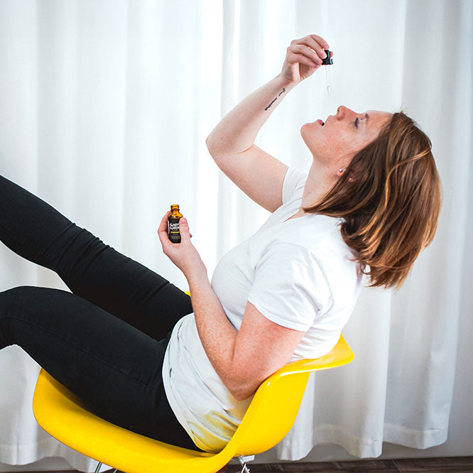 A woman tilts her head back on while sitting on a yellow chair and drops some CBD oil in her mouth