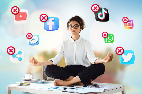 A woman meditates in lotus pose on her desk with her eyes closed while a bunch of x'd out social media apps fly around her