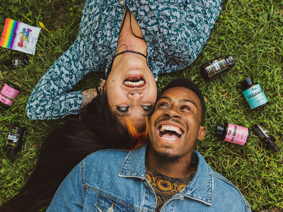 A man and a woman lie down on the grass smiling, surrounded by Sunday Scaries products