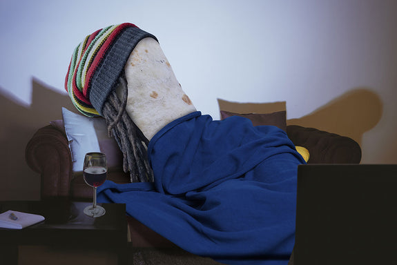 An actual burrito is wrapped in a blanket wearing a Rasta hat and drinking a glass of wine 