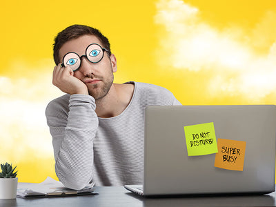 A man has fake glasses with painted eyes on them while he sleeps at work and pretends he's on his computer