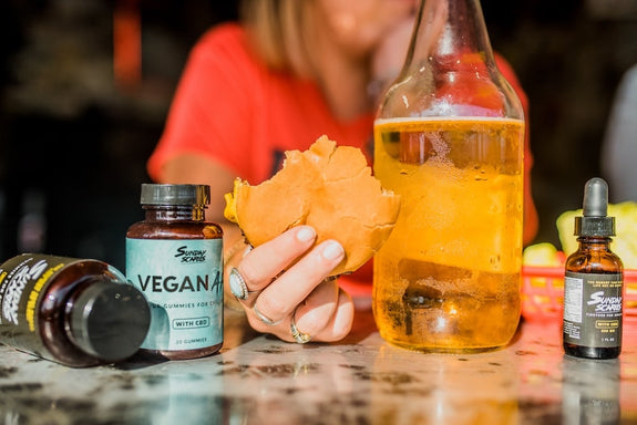 A woman holds a cheeseburger next to CBD gummies and a beer wondering if it's safe to mix CBD edibles and alcohol