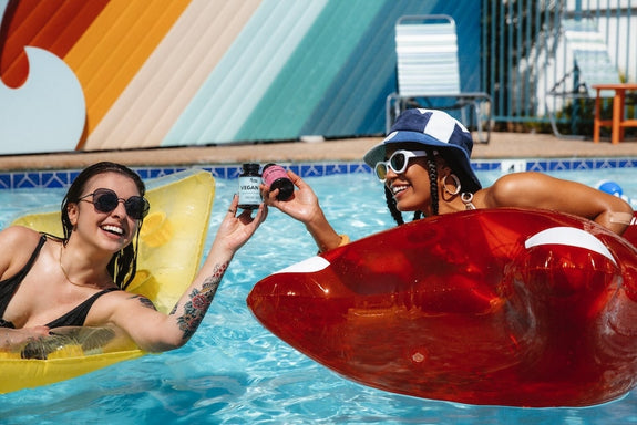 Two woman in a pool clank CBD gummies bottles in a pool to show if full spectrum vs broad spectrum is better for anxiety