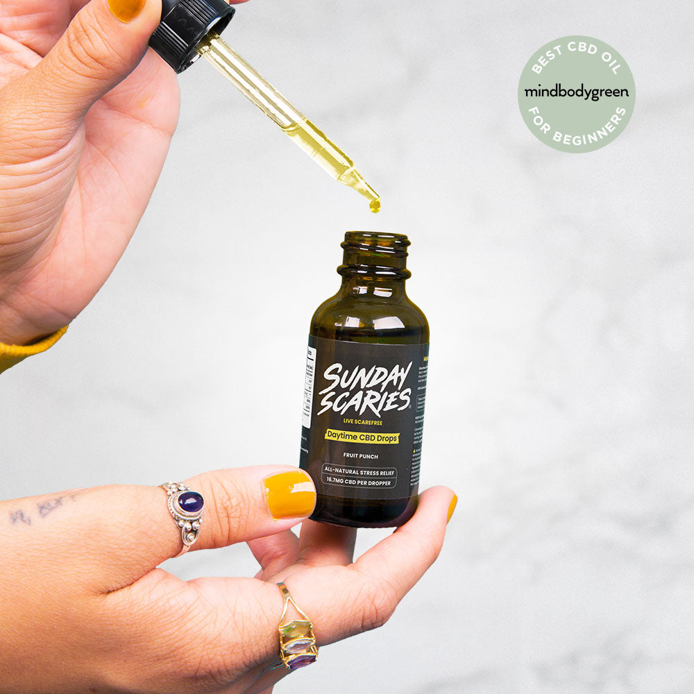 Hand holding a CBD oil bottle labeled 'Sunday Scaries' with a dropper, against a light gray background and a badge that says "Best CBD Oil for Beginners" from MindBodyGreen.