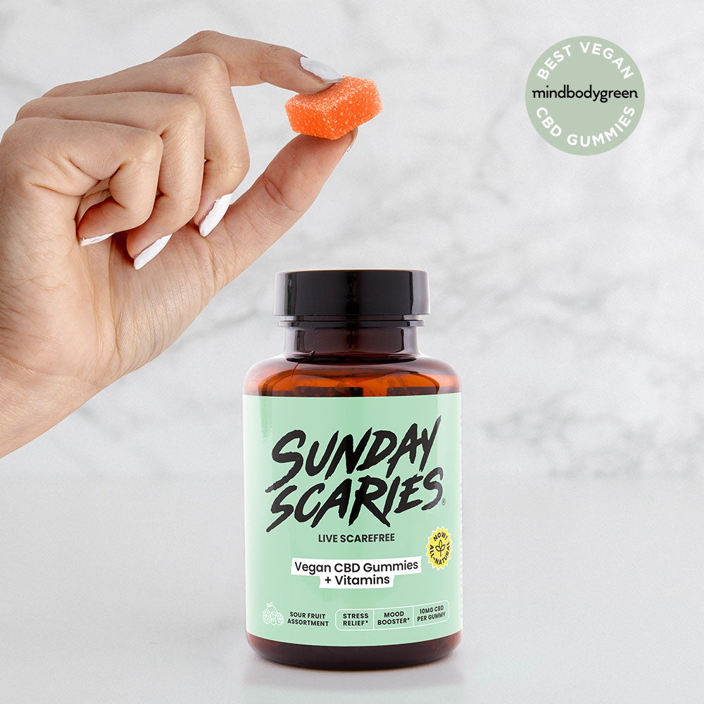 Hand holding a CBD gummy with a bottle labeled 'Sunday Scaries' on a light background and "Best Vegan CBD Gummies" badge from MindBodyGreen